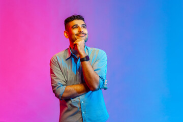 Portrait of man in shirt standing, touching his chin, toothy smile and looking away dreaming,...