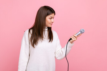 Side view portrait of brunette female offering microphone to somebody, journalist asking opinion, wearing white casual style sweater. Indoor studio shot isolated on pink background.