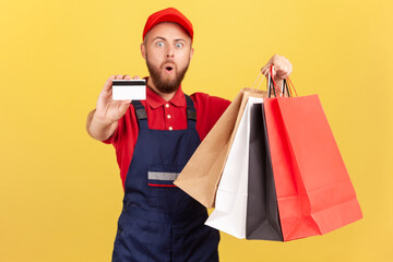 Portrait of surprised shocked deliveryman in blue uniform, red t-shirt and hat holding shopping bag and credit card, cashless payments. Indoor studio shot isolated on yellow background.