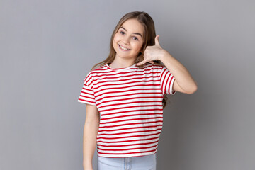 Obraz premium Portrait of satisfied little girl wearing striped T-shirt holding fingers near ear showing call gesture with toothy smile, answering call. Indoor studio shot isolated on gray background.