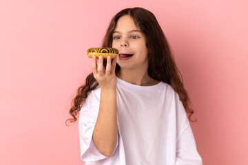 Portrait of delighted cute little girl wearing white T-shirt licking delicious donut, looking with desire to eat sweet dessert, showing tongue out. Indoor studio shot isolated on pink background.