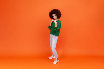 Full length portrait of woman with Afro hairstyle wearing green casual style sweater and jeans, standing with raised fists, expressing anger. Indoor studio shot isolated on orange background.