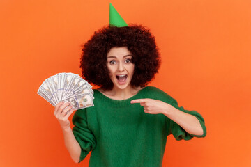 Excited woman with Afro hairstyle wearing green casual style sweater and party cone, having big sum of money as birthday present, pointing. Indoor studio shot isolated on orange background.