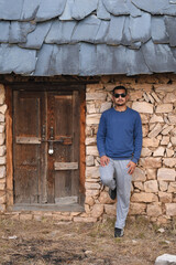 Man standing leaning against old structure wall made of stone and mud mixture looking straight wearing black sunglasses