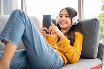 Beautiful asian young woman wearing headset and using mobile smartphone. Female leisure on sofa and enjoy watching device screen smart phone