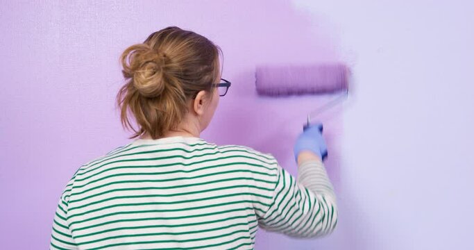 Young tired woman listlessly paints wall with paint roller in lilac color. Color therapy influence of color on human condition Lady indifferently paints the wall purple, she is tired, depressed.