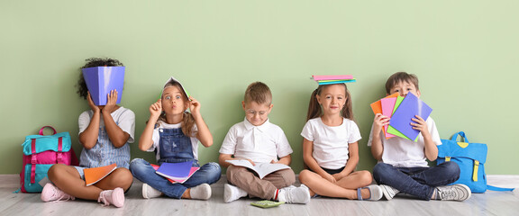 Funny little children with copybooks sitting on floor near green wall - obrazy, fototapety, plakaty