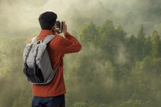 Hipster young asian man on the top of mountain with backpack in the green forest and take a photo with digital camera the beauty of landscape, Adventure and travel in the mountains region concept