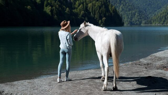 Mysterious woman tourist with animal horses wild white sunset running rider cowboy water Amazing view turquoise water high mountain lake Braies mountains. Casual boho style clothing back view