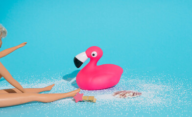 Plastic flamingo and girl doll, sand and seashell on cyan background