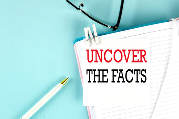 UNCOVER THE FACTS text on a sticky on notebook with pen and glasses , blue background
