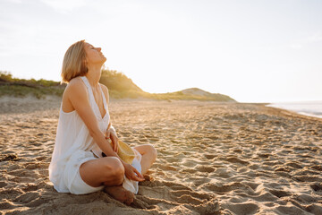 Side view of slim woman with short fair hair wearing white light summer sundress, sitting with crossed legs on sandy beach near water on sunny day, sunbathing. Summer, holiday, meditating, vacation.