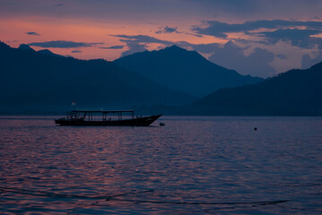 Tender sunset on ocean with gentle colorful pink heaven, clouds, calm smooth water with purple sun glare, blue mountains in haze, floating boat. Rich saturated indonesian sea landscape, trip in asia.