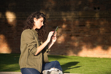 Beautiful young latina woman with curly brown hair and dressed in casual clothes sitting on a grey wall checking her mobile phone, social media, shopping and email. Technology concept.