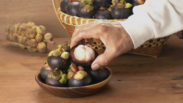 Man taking a half of peeled mangosteen in a wooden plate on wooden table with a bunch of longkong and a basket of mangosteen in the background.