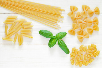 Pasta, set of raw farfalle, spaghetti, pipe, maccheroni, green leaf basil, on white wooden board background, top view, space to copy text.