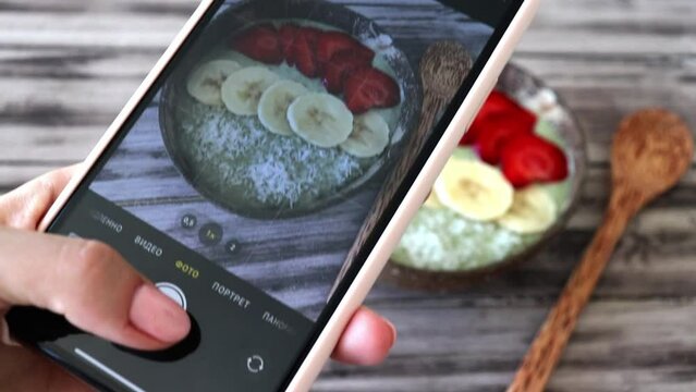 Woman takes a picture of her breakfast on her phone. Healthy superfood smoothie bowl with fruit berries and coconut shavings.