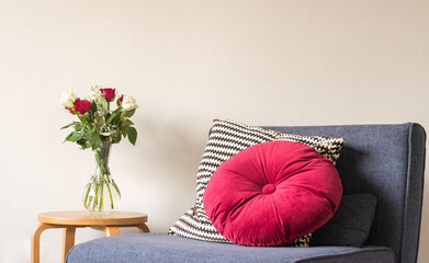 Red and cream roses in glass vase on small wooden table next to grey armchair with bright cushion  against beige wall (selective focus)