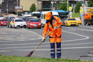 Woman worker in orange uniform with a broom sweeps the road on cars background. Traffic jam, street cleaning in summer city