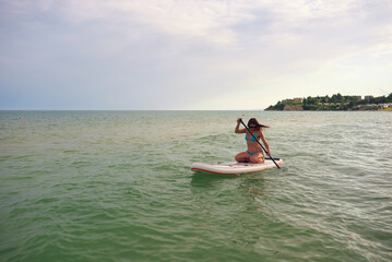 Woman in a blue bikini sits on a sup board with a paddle in the sea against the sunset.