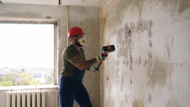 Contractor wrecks concrete wall with sledgehammer for rearrangement. Man doing manual dismantling and demolition works with big hammer hits for apartment renovation. Construction worker in uniform.