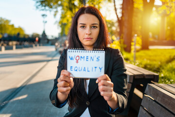 Young caucasian woman outdoors showing a notebook paper in front of her face with the text womens...