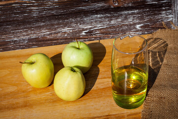 A glass of freshly squeezed apple juice and a green ripe apples on a table covered with a cloth against a wooden textured wall.