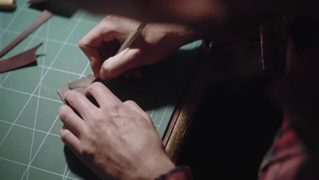 Close-up hands carve with the knife the pattern of leather and remove excess part. Tanner, tailor master works behind the workbench and creating leather goods. Handmade.