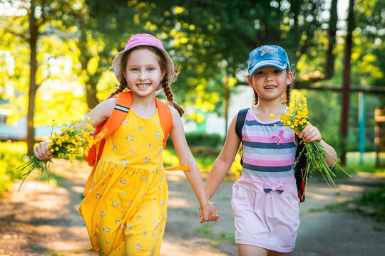 Two funny little girls of different races, in dresses and baseball caps, with a bouquet of wildflowers, run and laugh. Best friends like to chat and walk in the fresh air. Summer holidays