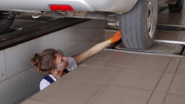 Female mechanic inspecting car suspension on hydraulic wheel play detector with remote control. Strong woman checks for wear in running gear and steering components. Service operator in overalls.