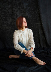 portrait of sad beautiful woman in white shirt and blue jeans sitting in studio on black background