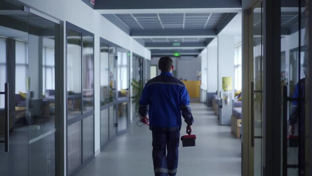 Electrician goes to repair light in office. Master finishes work with electricity. Locksmith walking along business center hall with toolbox. Male mechanic in uniform.