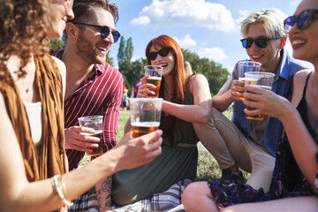 Group of young caucasian friends sitting on grass, drinking beer and  having fun on music festival
