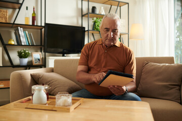 Senior man drinking tea and reading news article on tablet computer at home