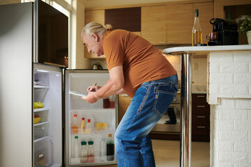 Elderly man looking at opened fridge and writing down what to buy in grocery store