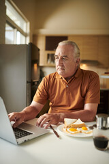 Fototapeta na wymiar Elderly man checking his social media account on laptop when eating breakfast at home