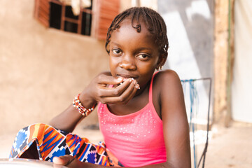 Close-up of a cute African girl sitting in a courtyard eating rice with her right hand; human...