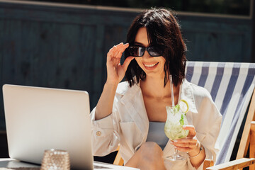 Cheerful Young Woman in Sunglasses Drinking Cocktail and Using Laptop for Video Call While Sitting on Terrace