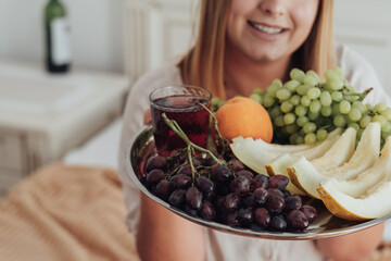 Unrecognisable Young Woman Holding Tray of Fruit in Hotel Room, Bottle of Wine Stands on the Background