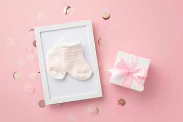 Baby concept. Top view photo of white giftbox with ribbon bow photo frame with tiny socks and shiny confetti on isolated pastel pink background