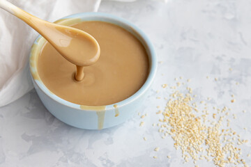 Close up of traditional sause tahini in spoon over ceramic bowl and sesame seeds on light grey background. Selective focus. Blurred background.