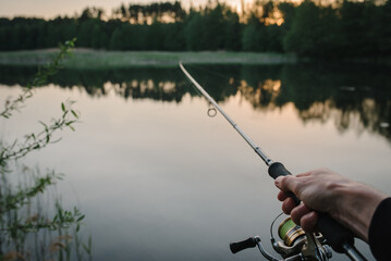 Fisherman with rod, spinning reel on the river bank. Man catching fish, pulling rod while fishing from lake or pond with text space. Fishing for pike, perch on beach lake or pond. Fishing day concept.