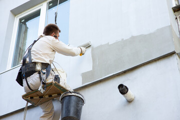 Builder-alpinist paints the facade of the building in a light color - obrazy, fototapety, plakaty