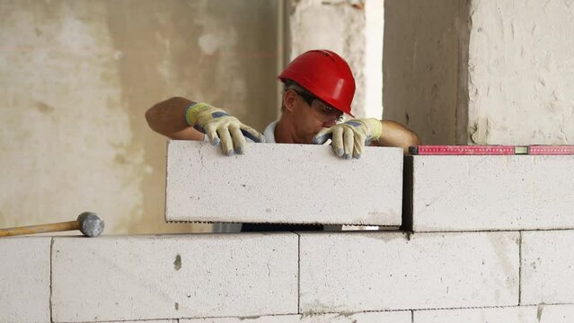 Bricklayer or mason lays bricks to construct wall of autoclaved aerated concrete blocks. Brickwork worker contractor constructs blockwork with foam concrete doing masonry on construction site.