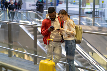 Young couple of travelers using smartphones while waiting for flight at airport. Two multiracial...