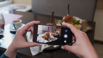 A woman takes a picture on a smartphone of a delicious burger on a table in a cafe for social networks.