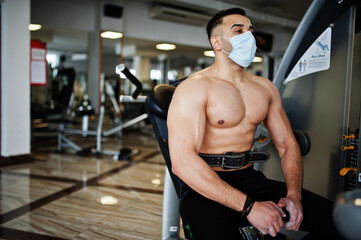 Muscular arab man, wear medical face mask training in modern gym. Fitness arabian men with naked torso doing exercises, durig coronavirus pandemic.