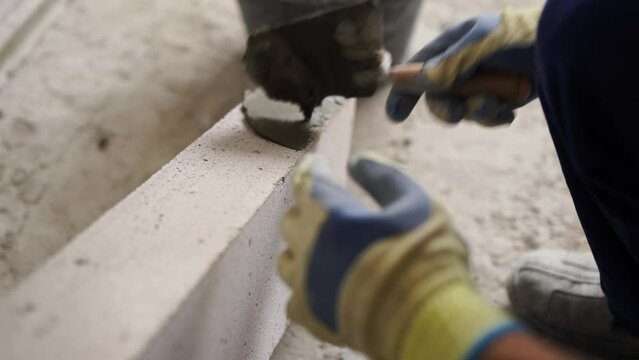 Bricklayer applies adhesive glue on autoclaved aerated concrete blocks with trowel and spatula. Brickwork worker contractor puts mortar on foam concrete doing masonry on construction site.