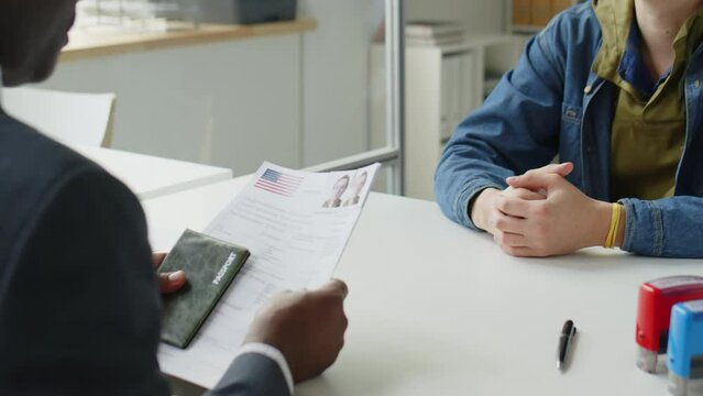 Tilt up shot of young Caucasian applicant giving documents to consulate officer and answering his questions during visa interview in U.S. embassy