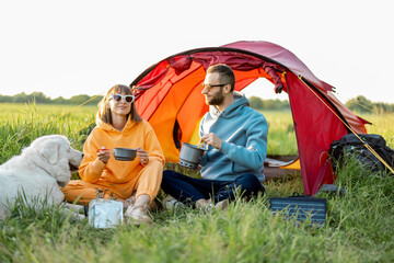 Young couple cooking food, spending summer time with a dog at campsite on the green field. Man and woman travel with tent on nature